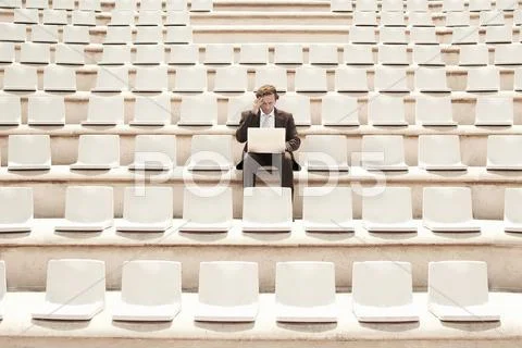 Confused Businessman Working On Laptop In Center Of Empty Auditorium Stock Photos