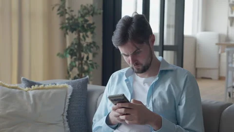 Confused Man Reading Unexpected Message On Smartphone While Sitting On Couch Stock Footage 303085940