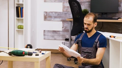 Confused worker rubbing his head while reading instruction for new furniture Stock Footage 123344917