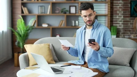 Confused young man having difficulty with paperwork while using mobile  Stock Footage 296580457
