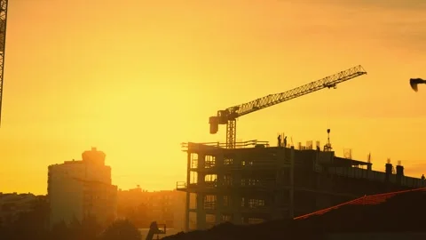 Construction crane rises above a building frame at sunset during work on a city Stock Footage 348295381