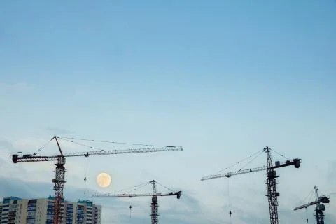 Construction cranes on the background of a clear sky and a large moon. Stock Photos