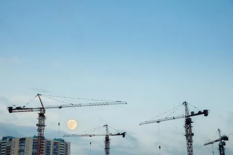 Construction cranes on the background of a clear sky and a large moon. Stock Photos