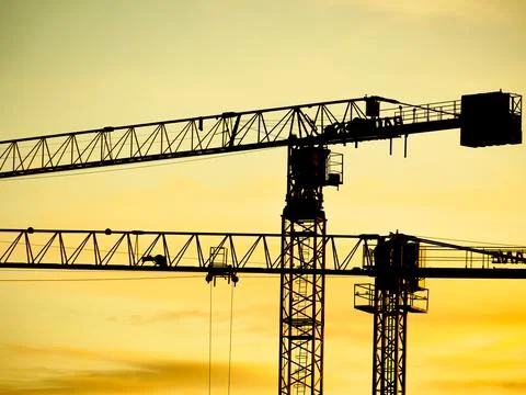 Construction cranes stand against a sunset sky in an urban area with clouds i Stock Photos