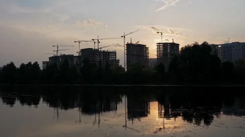 Construction Cranes Towering Over Urban Skyline Near a Calm Lake at Sunset Stock Footage 345555224