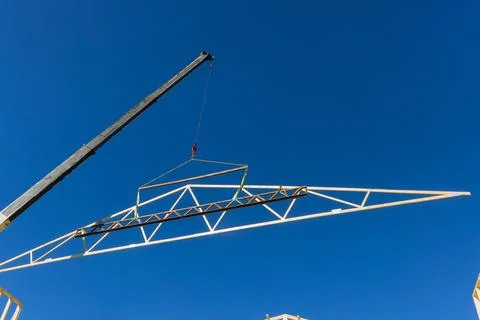 Construction crew lifts an engineered truss using crane on clear sunny day in Stock Photos