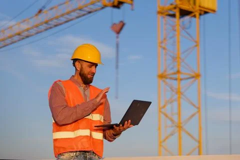 Construction engineer using laptop at building site with crane in background Photos