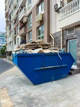 Construction materials and waste are piled high in a blue dumpster on the sid Stock Photos
