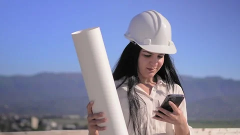 Construction site in helmet holds mobile phone and rolled-up paper plan 库存影片 240340401