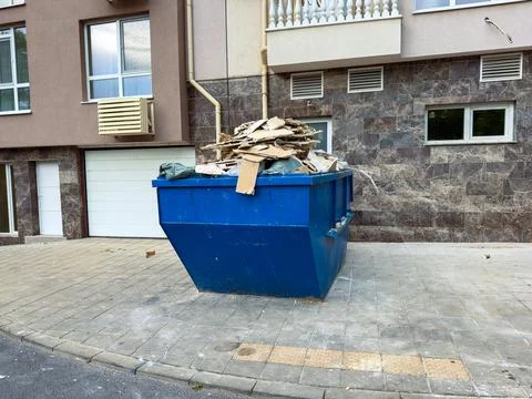 Construction site waste is piled in a blue dumpster near a modern building .. Stock Photos