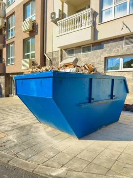 Construction waste is piled in a blue dumpster outside a building on a sunn.. Stock Photos