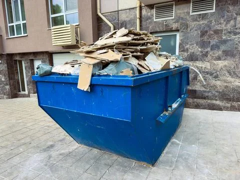 Construction waste is piled high in a blue dumpster located near a building.. Stock Photos