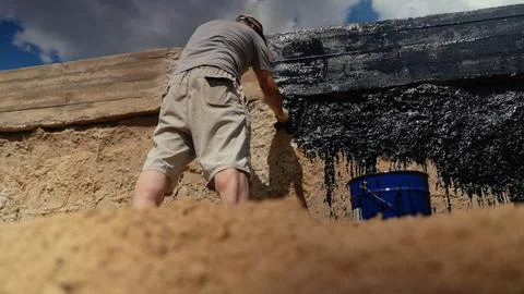 Construction worker applying bitumen waterproofing to building foundation Stock Photos