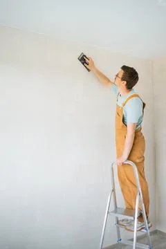 Construction worker in beige overalls balancing on metal ladder, sanding white Stock Photos
