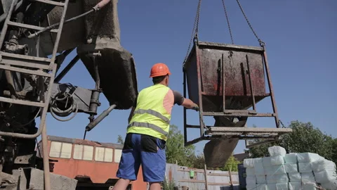 Construction worker guides concrete hopper suspended beside mixer truck at Видео 343409889