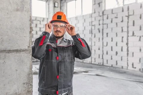 Construction worker in a hard hat and overalls adjusts his safety glasses at a Stock Photos