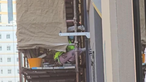 A construction worker in a high-visibility vest hammers on scaffolding 스톡 동영상 311885763