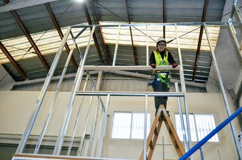 Construction worker mounting plasterboard on metal frame using wooden ladder Stock Photos