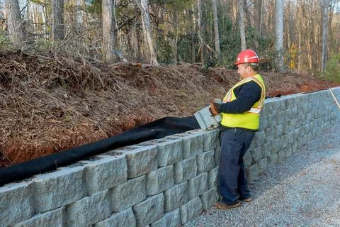 Construction worker placing stone blocks for retaining wall along forested road Stock Photos