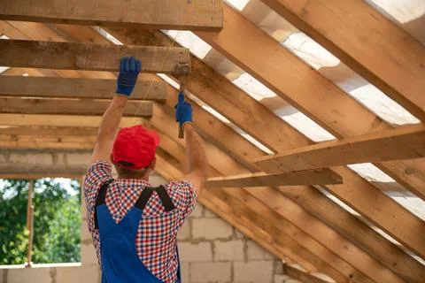 Construction worker in red cap and blue overalls installing wooden roof tru.. Stock Photos