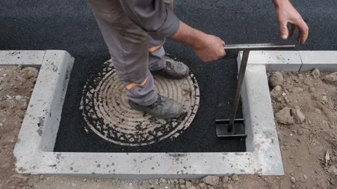 Construction worker tamping fresh hot asphalt around a manhole cover using .. Stock Footage 339802581