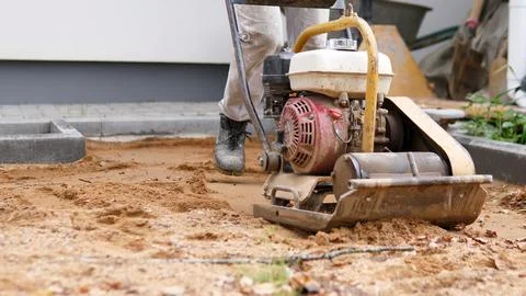 Construction worker using plate compactor on sand beside concrete curb during Stockfoto's