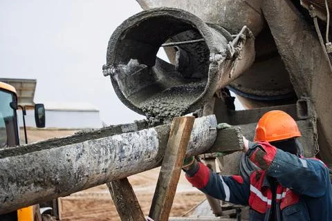 A construction worker wearing a hard hat pours wet concrete from a mixer into Stock Photos