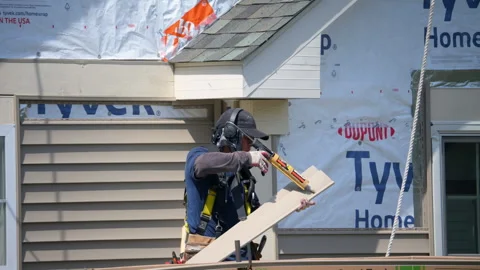 Construction worker wearing safety harness installs exterior wall siding on a Stock-Footage 328072666