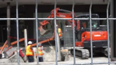 Construction workers and an orange excavator behind a wire fence at an urban Vidéo 334583272