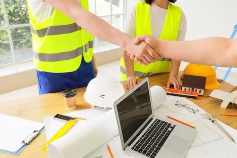 Construction workers in safety vests shaking hands over blueprints and laptop Stock Photos