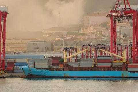 Container ship and red cranes at Lisbon port with cityscape and fire smoke in Stock Photos