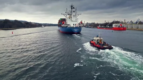 A container ship escorted by a red tugboat in a narrow waterway near a port Stock Footage 300257401