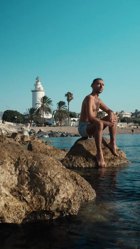 Contemplating man with man bun gazing at distant lighthouse on rocky coast,.. Stock Footage 344059264