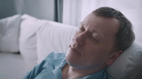 A contemplative man is sitting in soft light on a cozy couch, embodying a Stock Footage 316619134