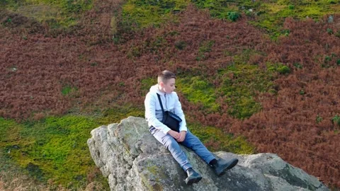 A contemplative young boy rests on a sizable rock outcrop amidst the moorla.. Видео 254160359
