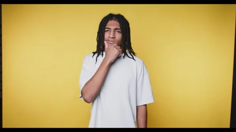 Contemplative young man with braids holds his chin, looking thoughtfully at.. Stock Footage 313280103