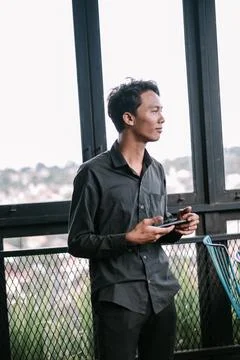 Contemplative young man using cellphone by window overlooking the city Stock Photos