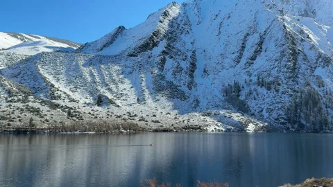 Convict Lake with a tiny kayak crossing mirror-like water below snow covered Video stock 324712982