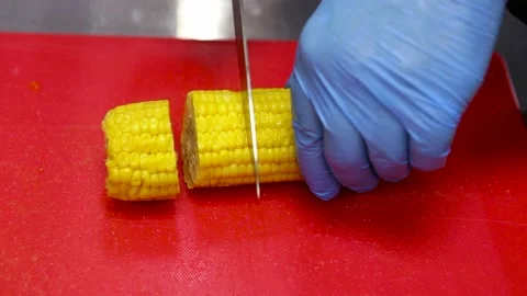 Cook in blue gloves cuts corn cobs on a red chopping board. Close-up Stock Footage 130903752