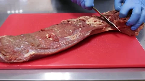 Cook in blue gloves cuts raw meat on a red chopping board. Close-up Stock Footage 130903736