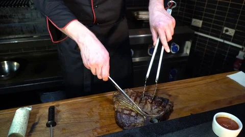 The cook cuts the steak with a kitchen knife and tongs in the kitchen Stock-Footage 85671472