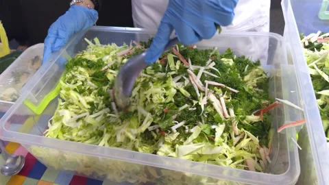 Cook in gloves adding cucumbers to a large cabbage salad in a canteen kitchen Video stock 333622340
