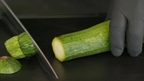 A cook slices a courgette lengthwise with a knife on a black board.  Close up Stock Footage 163044572