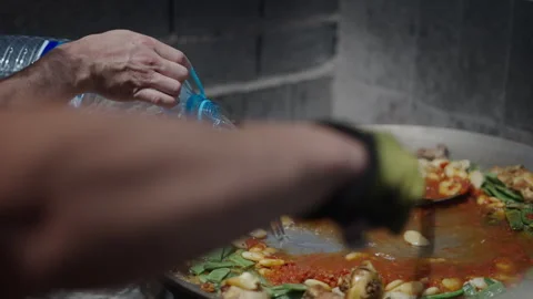 The cook stirs the paella while the helper pours water Stock Footage 259958010