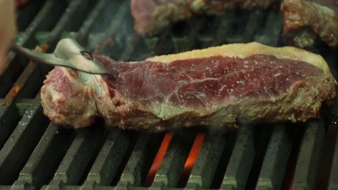 The cook turns tall steaks with a fork on a flaming smoking hot char broiler  Stock Footage 127715329