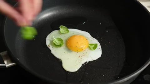 Cooking fried eggs in frying pan. Hands garnish egg with fresh basil leaves Stock Footage 340330934
