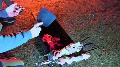 Cooking meat over an open fire at a campsite during a night gathering with frien Stock Footage 326116144