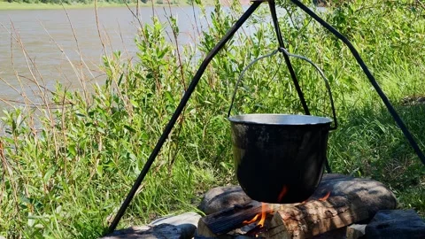 Cooking over fire by the river. The pot hangs over fire, flames dance around it Stock Footage 279558791