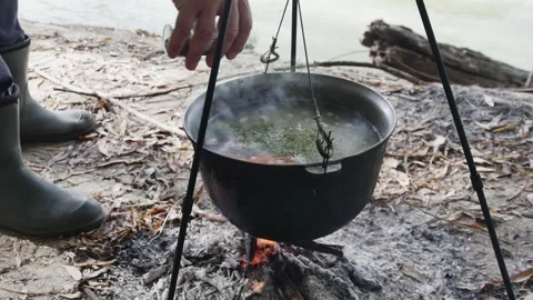 Cooking river fish soup in a cauldron on a fire on the river bank, a fisherm Vidéo 254061844