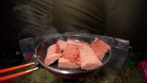 Cooking spam processed meat in a frying pan inside a dark windy camping tent Stock Footage 284024212